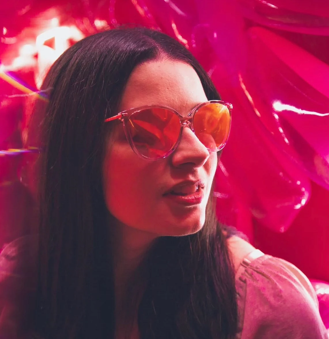 Portrait of a woman in rose-tinted sunglasses lit by magenta light bouncing off crumpled foil, her face half in shadow.