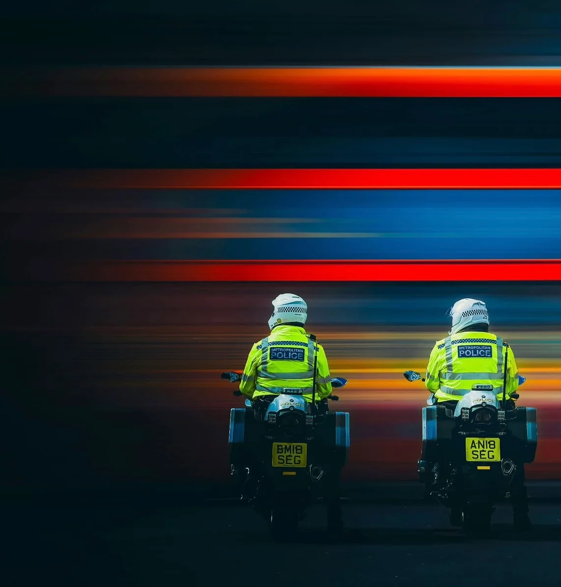Two police motorcyclists in high-visibility jackets held still against a wall of red and blue light trails on a London street at night.