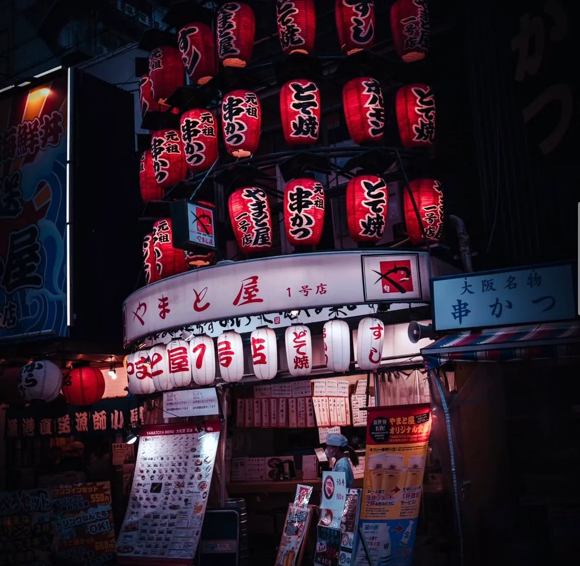 A kushikatsu shopfront in Shinsekai stacked with dozens of red paper lanterns, a lone cook working under the glow.